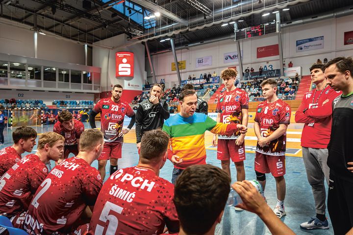 Eine Handballmannschaft steht in einer Sporthalle  im Kreis um ihren Trainer herum. 