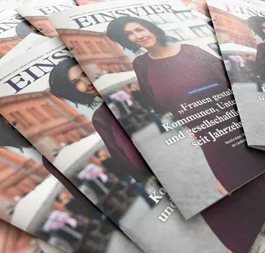 EINSVIER 1-2026 Mehrere Magazine liegen aufgefächert auf einem Tisch. Auf dem Titelbild ist eine Frau mit dunklen Haaren und einem lilafarbenen Kleid zu sehen.