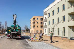 Zu sehen ist eine Baustelle mit fast fertiggestellten Häusern. Im Vordergrund pflanzen zwei Männer einen Baum an eine Straße. Links steht ein Arbeitsfahrzeug.