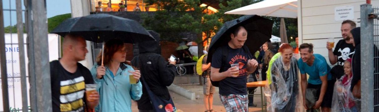 Menschen mit Regenschirmen und weitere Personen, die sich unter einem Dach unterstellen.