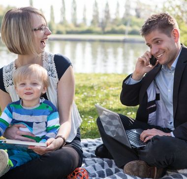 Eine Familie sitzt zusammen am Wasser, der Vater arbeitet mit dem Mobiltelefon