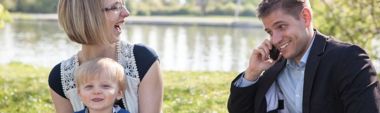 Eine Familie sitzt zusammen am Wasser, der Vater arbeitet mit dem Mobiltelefon