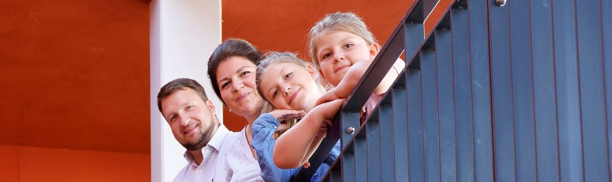 Eine Familie mit zwei Kindern blickt über eine Treppenbrüstung nach unten in die Kamera.