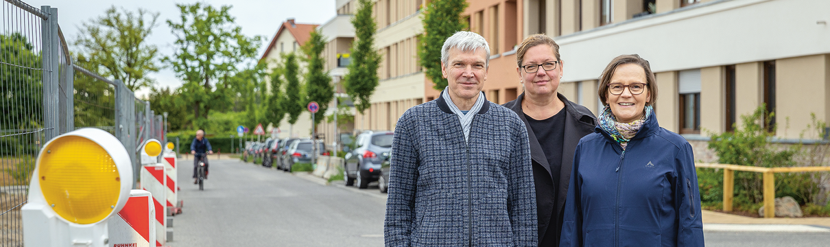 Zwei Frauen und ein Mann stehen auf einer Straße. Auf der rechten Seite sind im Hintergrund Wohnhäuser, links ein Bauzaun. 