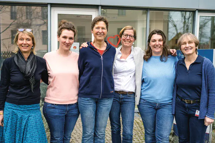Zu sehen sind sechs Frauen, die nebeneinander stehen, die Arme umeinandergelegt haben und in die Kamera lächeln.