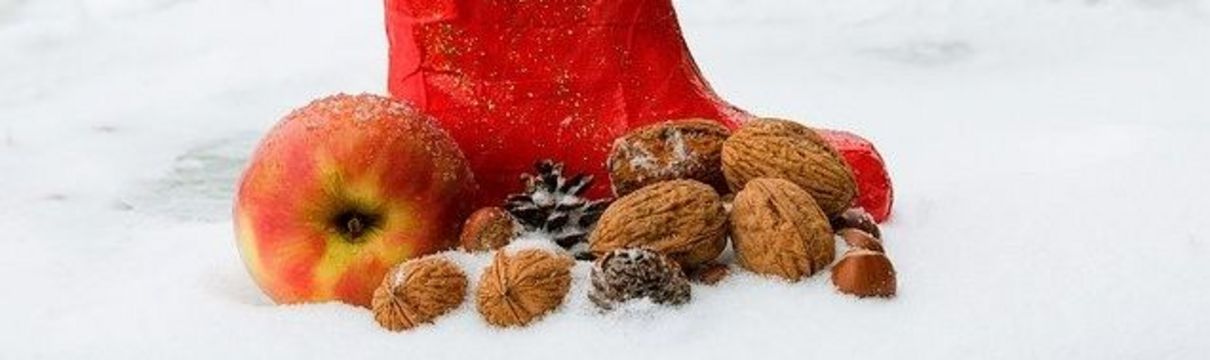 Zu sehen ist ein roter Nikolausstiefel, der im Schnee steht und gefüllt und umgeben ist von Nüssen, Tannenzapfen und einem Apfel.