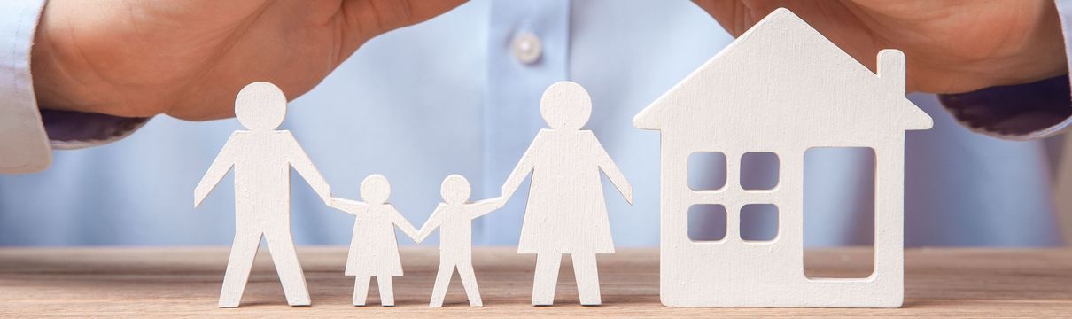 Weißer Scherenschnitt aus vierköpfiger Familie neben einem Haus, im Hintergrund steht eine reale Person, die ihre Hände schützend darüber hält