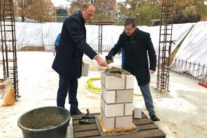 Der Geschäftsführer der ProPotsdam und der Oberbürgermeister der Stadt Potsdam tragen mit Schippen eine graue Masse auf einen Stapel mit steinernen Bauziegeln auf.