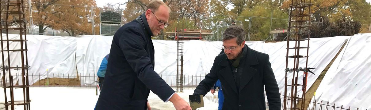 Der Geschäftsführer der ProPotsdam und der Oberbürgermeister der Stadt Potsdam tragen mit Schippen eine graue Masse auf einen Stapel mit steinernen Bauziegeln auf.