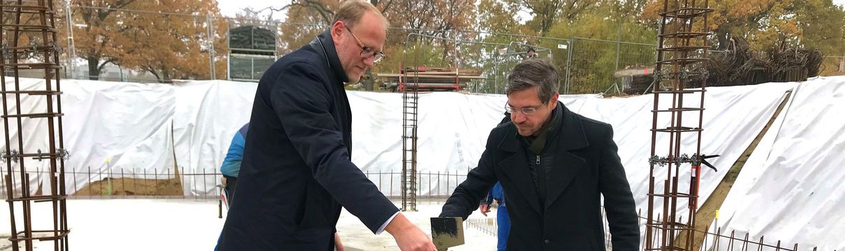Der Geschäftsführer der ProPotsdam und der Oberbürgermeister der Stadt Potsdam tragen mit Schippen eine graue Masse auf einen Stapel mit steinernen Bauziegeln auf.
