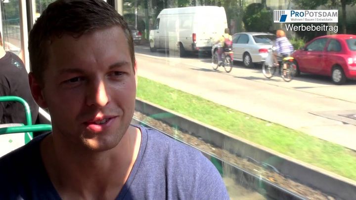 Ein männlicher Fahrgast mit blauem T-Shirt sitzt in einer fahrenden Straßenbahn. Man sieht durch die Fnesterscheibe der Bahn einen Teil der Schienen und den angrenzenden schmalen Grünstreifen, der eine einspurige Straße und seitlich parkende Autos von den Schienen trennt.