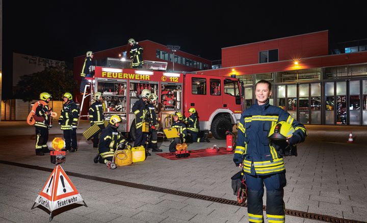 Eine junge Frau in Feuerwehrausrüstung steht bei Nacht im Vordergrund, im Hintergrund ein Feuerwehrfahrzeug mit mehreren Feuerwehrmännern.