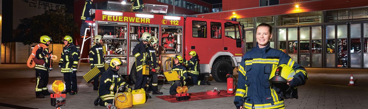 Eine junge Frau in Feuerwehrausrüstung steht bei Nacht im Vordergrund, im Hintergrund ein Feuerwehrfahrzeug mit mehreren Feuerwehrmännern.