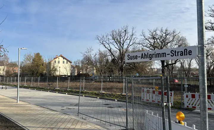 Straßenschild „Suse-Ahlgrimm-Straße“ an einer ruhigen Straße mit Gehweg, Bauzaun und Wohnhäusern im Hintergrund.