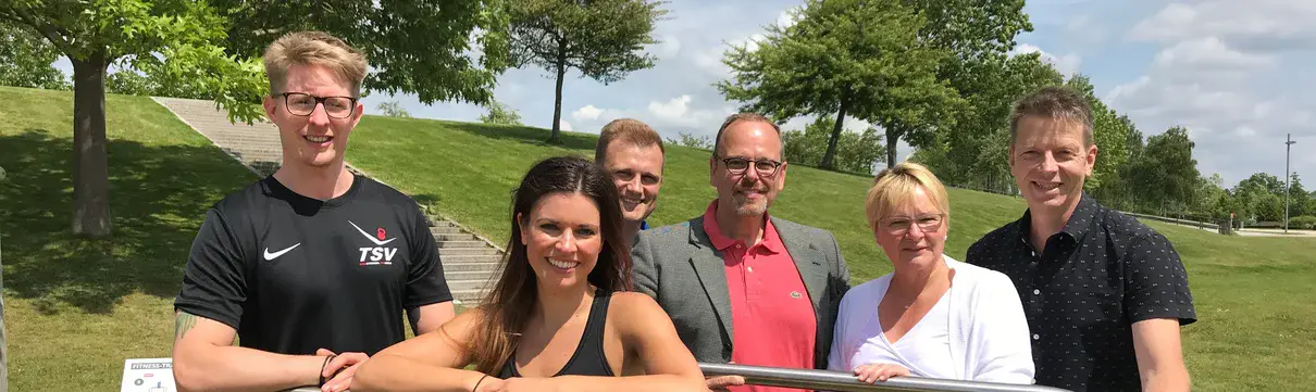 Eine Gruppe von Menschen steht an einem metallenem Bügel. Zwei der Personen tragen Sportkleidung.