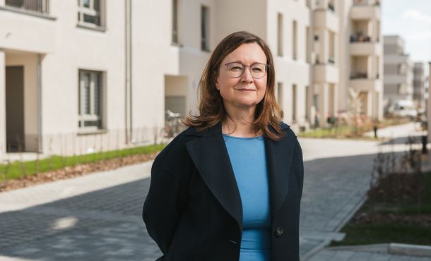 Porträt einer Frau mit braunen, halblangen Haaren, Brille, blauem Oberteil und schwarzem Sacko vor einer sonnigen Häuserzeile stehend