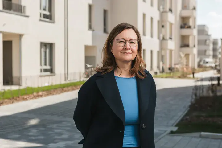 Sandra Jacob Porträt einer Frau mit braunen, halblangen Haaren, Brille, blauem Oberteil und schwarzem Sacko vor einer sonnigen Häuserzeile stehend
