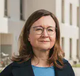 Porträt einer Frau mit braunen, halblangen Haaren, Brille, blauem Oberteil und schwarzem Sacko vor einer sonnigen Häuserzeile stehend