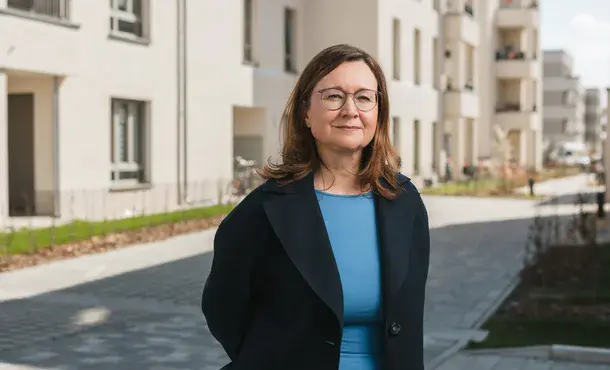Porträt einer Frau mit braunen, halblangen Haaren, Brille, blauem Oberteil und schwarzem Sacko vor einer sonnigen Häuserzeile stehend