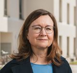 Porträt einer Frau mit braunen, halblangen Haaren, Brille, blauem Oberteil und schwarzem Sacko vor einer sonnigen Häuserzeile stehend