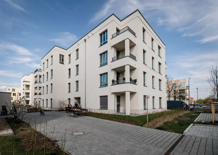 Modernes, helles Mehrfamilienhaus mit vier Stockwerken und Balkonen in einem neu angelegten Wohnquartier mit Gehwegen und blauem Himmel