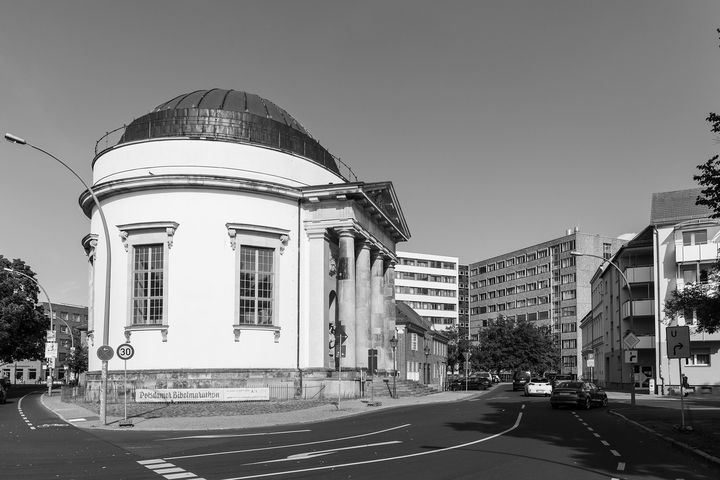 Schwarz-Weiß-Foto eines Pantheons direkt auf der Ecke einer Kreuzung. Im Hintergrund ragt ein mehretagiger Plattenbau empor.