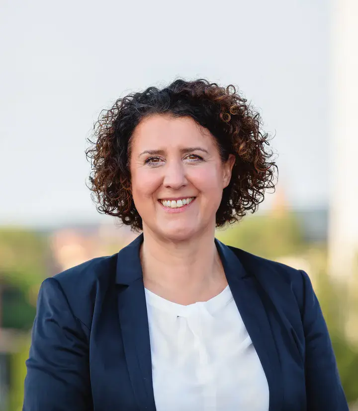 Portraitbild einer dunkelhaarigen Frau, die lächelt, im Hintergrund Gebäude