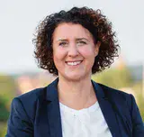 Portraitbild einer dunkelhaarigen Frau, die lächelt, im Hintergrund Gebäude