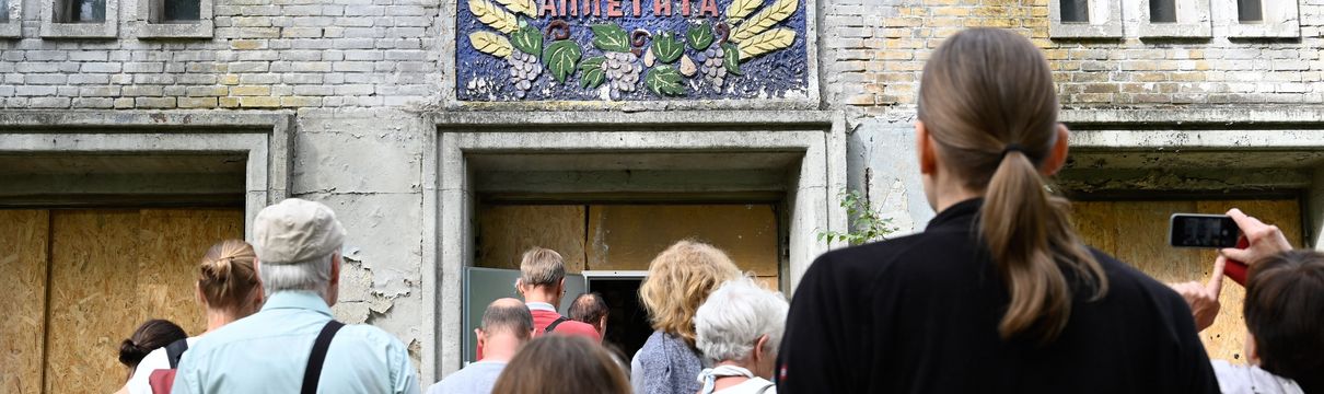 Ein Gruppe von Menschen steht mit dem Rücken zur Kamera vor einem leicht verfallenen Gebäude mit einer russischen Inschrift über der Tür.