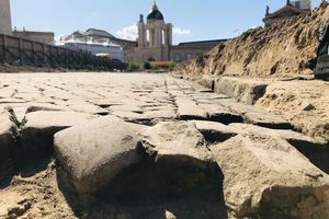 Straßenplaster, das am unteren Bildrand aufgebrochen ist. Rechts zieht sich ein Sandhügel entlang.