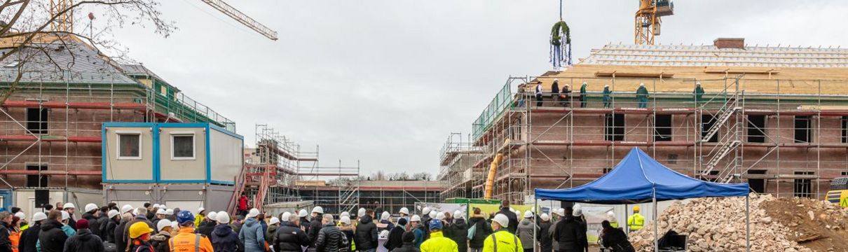 Zu sehen sind Menschen mit weißen Bauhelmen auf den Köpfen, die auf einer Baustelle vor einemengerüsteten Gebäude stehen.