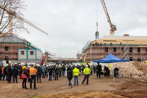 Zu sehen sind Menschen mit weißen Bauhelmen auf den Köpfen, die auf einer Baustelle vor einemengerüsteten Gebäude stehen.