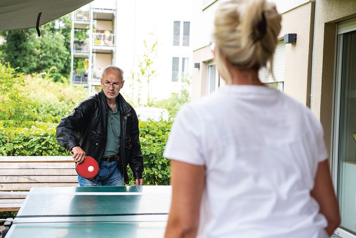 Ein Mann mit Brille in Lederjacke spielt Tischtennis mit einer blonden Frau im weißen T-Shirt, die nur von hinten zu sehen ist.