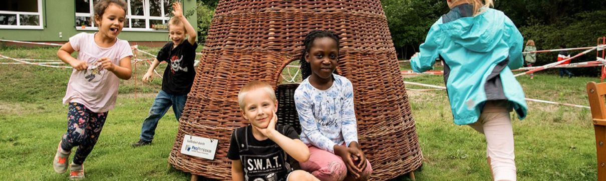 Mehrere Kinde spielen im Garten einer Kita um ein Zelt aus geflochtenen Zweigen