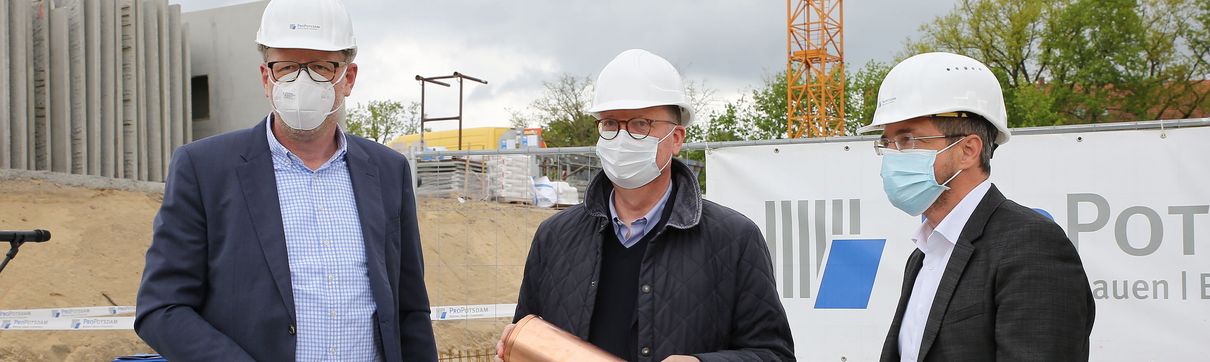Der ProPotsdam-Geschäftsführer, ein Architekt und der Oberbürgermeister stehen auf einer Baustelle. Alle tragen wei0e Schutzhelme. Der Architekt hält eine Zeitkapsel in der Hand. Im Hintergrund sind ein Banner der ProPotsdam und ein Kran zu sehen.