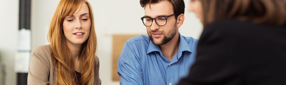Eine Frau und ein Mann schauen gemeinsam mit einer Beraterin auf etwas auf einem Tisch