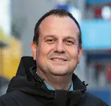 Portrait Tino Schröder, Teamleiter bei der ProPotsdam, lächelt in die Kamera. Im Hintergrund erstreckt sich die Konrad-wolf-Allee mit seinen bunten Balkonen.