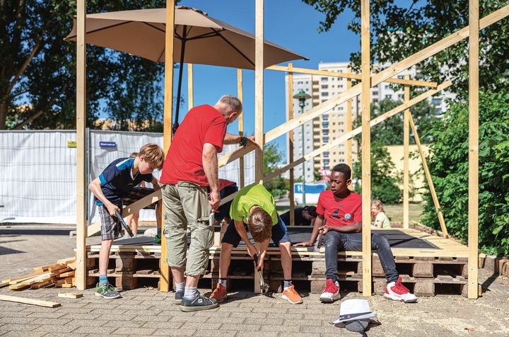 Drei Jungs bauen mit Hilfe eines Erwachsenen bei sonnigem Wetter eine Konstruktion aus Holzpaletten und Holzbrettern. Im Hintergrund sind Hochhäuser zu sehen.