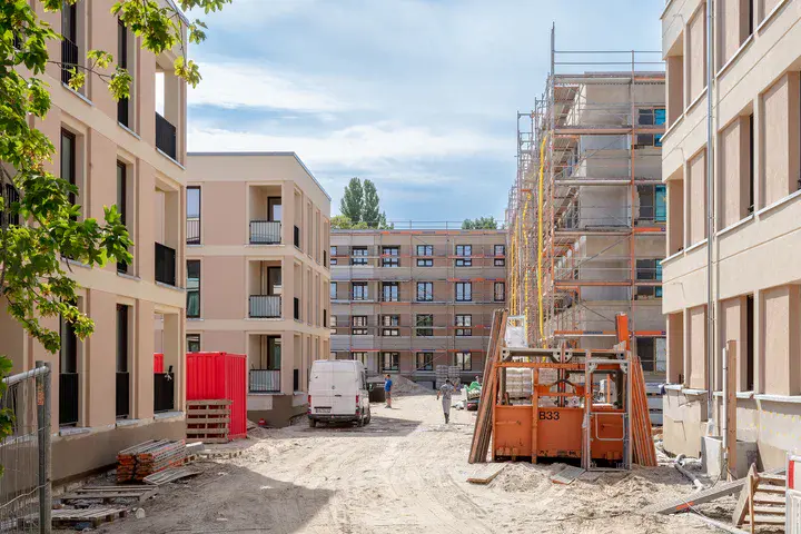 Auf einer Baustelle sind mehrere Gebäude, Baumaterialien und zwei Bauarbeiter zu sehen.