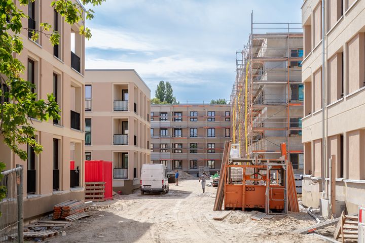 Auf einer Baustelle sind mehrere Gebäude, Baumaterialien und zwei Bauarbeiter zu sehen. 