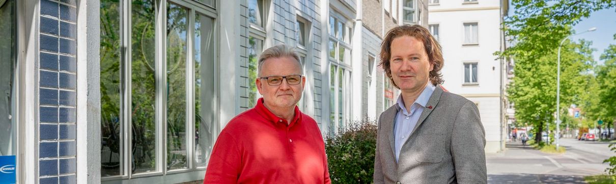 Ein Mann in einem roten Poloshirt und Mann in einem grauen Anzug stehen vor einer Häuserfassade