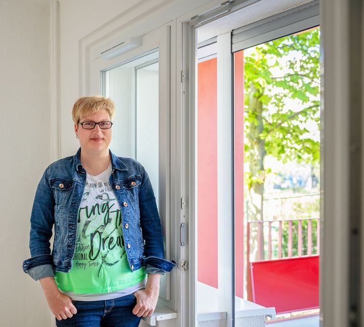 Eine Frau in Jeansjacke steht an einem Fenster in einer Wohnung