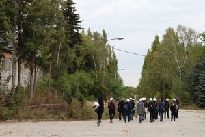 Menschen mit weißen Helmen gehen auf einem Weg durch ein mit Absperrbändern gesichertes Gelände mit alten Häusern und Bäumen..