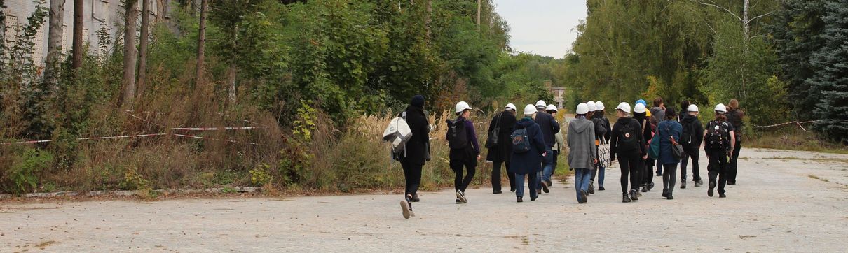 Menschen mit weißen Helmen gehen auf einem Weg durch ein mit Absperrbändern gesichertes Gelände mit alten Häusern und Bäumen..