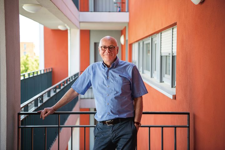 Älterer Mann mit Brille im blauen, kurzärmeligen Hemd steht lächeln in einem offenen Hausflur eines Wohnkomplexes mit oranger Hauswand