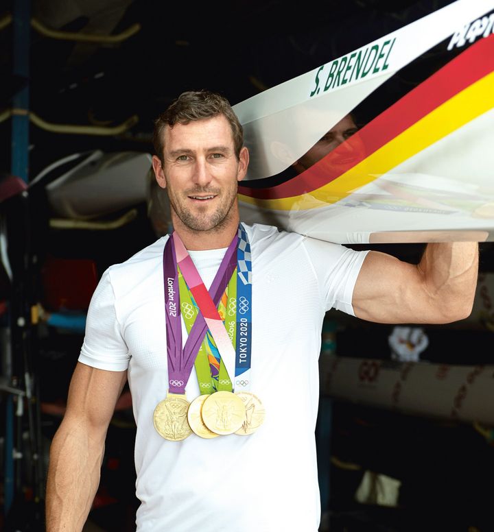 Ein lächelnder, sportlicher Mann mit vielen Medaillen um den Hals tragt ein Canadier-Sportboot auf der Schulter.