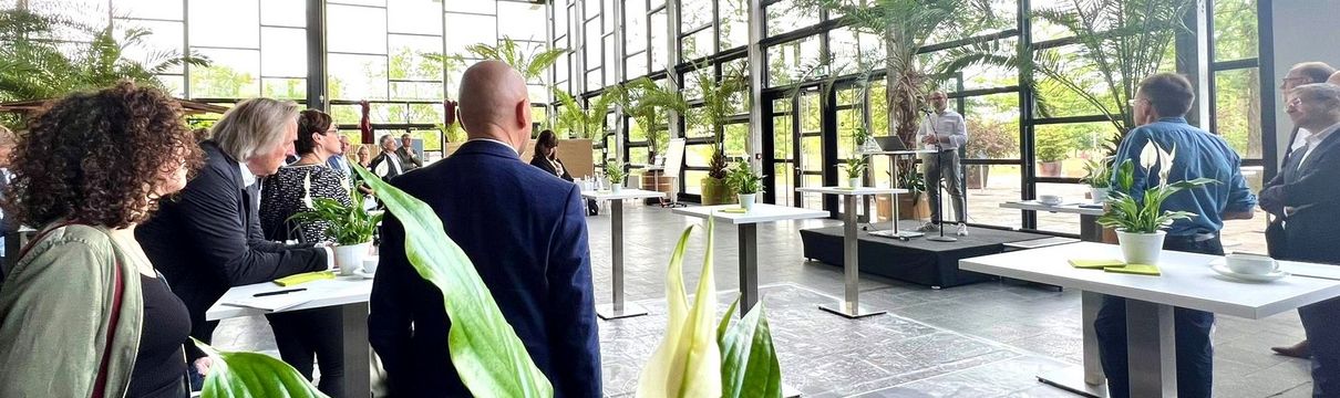 Mehrere Menschen stehen in der Biosphäre Potsdam an kleinen Tischen und hören ProPotsdam-Geschäftsführer Bert Nicke zu, der auf einer kleinen Bühne vor einem Mikrofon steht.  