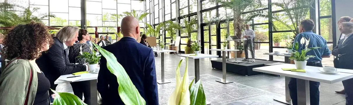Holzbausymposium Biosphäre Potsdam (Foto: Janine Klaus) Mehrere Menschen stehen in der Biosphäre Potsdam an kleinen Tischen und hören ProPotsdam-Geschäftsführer Bert Nicke zu, der auf einer kleinen Bühne vor einem Mikrofon steht.