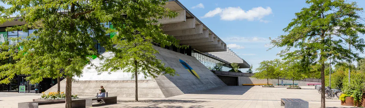 pyramidenartig gebautes Gebäude mit großem Flachdach, vor dem grüne Bäume stehen