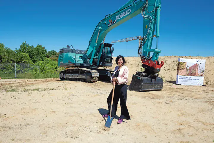 Potsdams Oberbürgermeisterin Noosha Aubel Eine Frau mit dunklen Haaren und hellem Blazer steht, auf einen Spaten gestützt, auf einer Baustelle. Im Hintergrund steht ein großer Bagger.